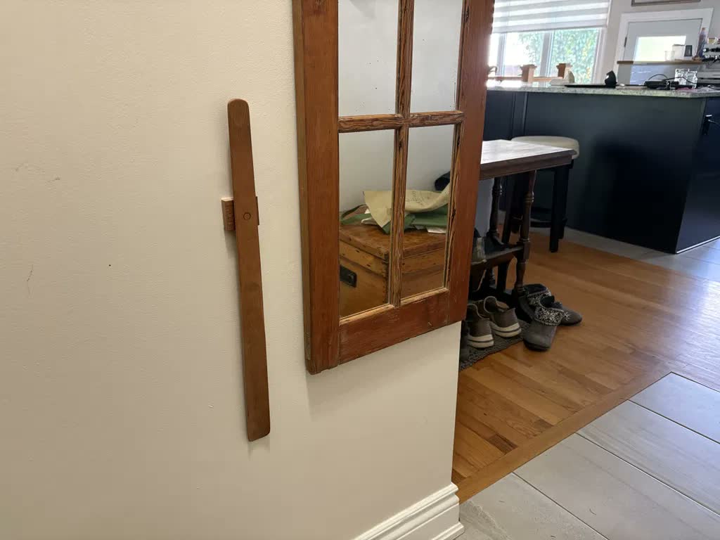 Calzascarpe in rovere chiaro su una parete crema accanto a un vecchio specchio con cornice in legno grezzo, con vista sulla cucina