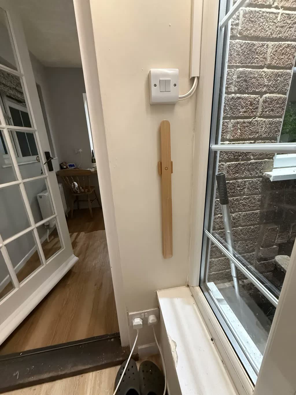 Calzascarpe in rovere chiaro su una parete crema vicino a una finestra e a una porta, con un interruttore sopra e degli zoccoli sul pavimento